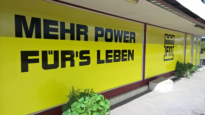Das Bild zeigt eine Werbefläche mit dem fehlerhaften Slogan "Mehr Power für’s Leben", der durch den Deppenapostroph bei für’s ein typisches Beispiel darstellt.